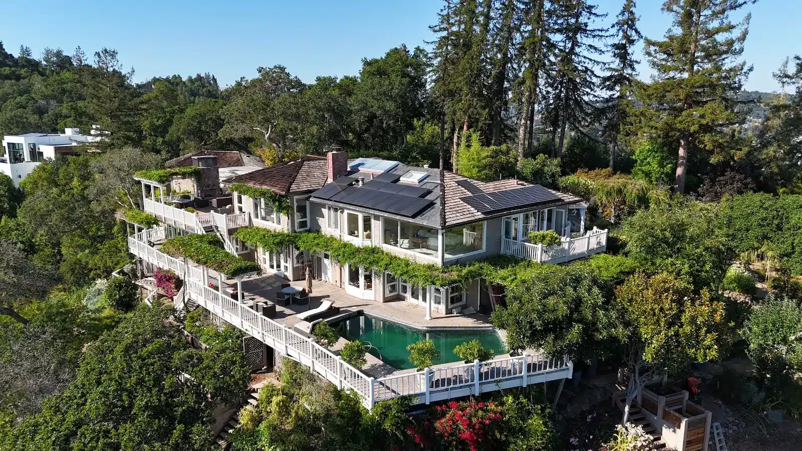 A large, luxurious house with multiple balconies, solar panels on the roof, and a pool surrounded by lush trees and greenery on a hillside under a clear blue sky.